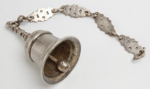 Campanilla de plata con vástago abalaustrado rematado en anilla. Se suspende de una cadena formada por cuatro eslabones laminares calados, que alternan con cinco eslabones de ese. Datada en la primera mitad del XVIII.